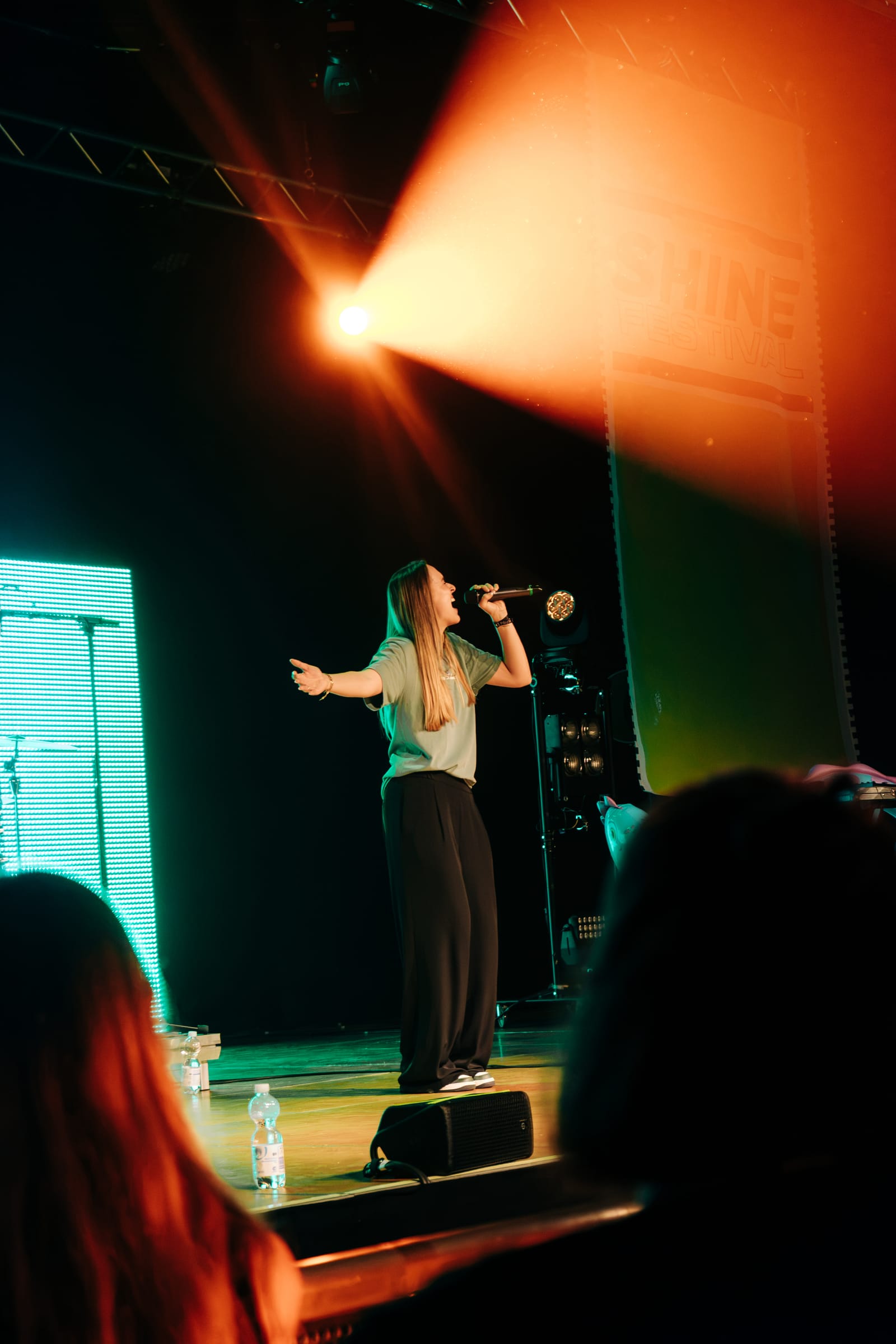 Toni Speer mid-performance, arms open, lit by a single orange stage lamp.